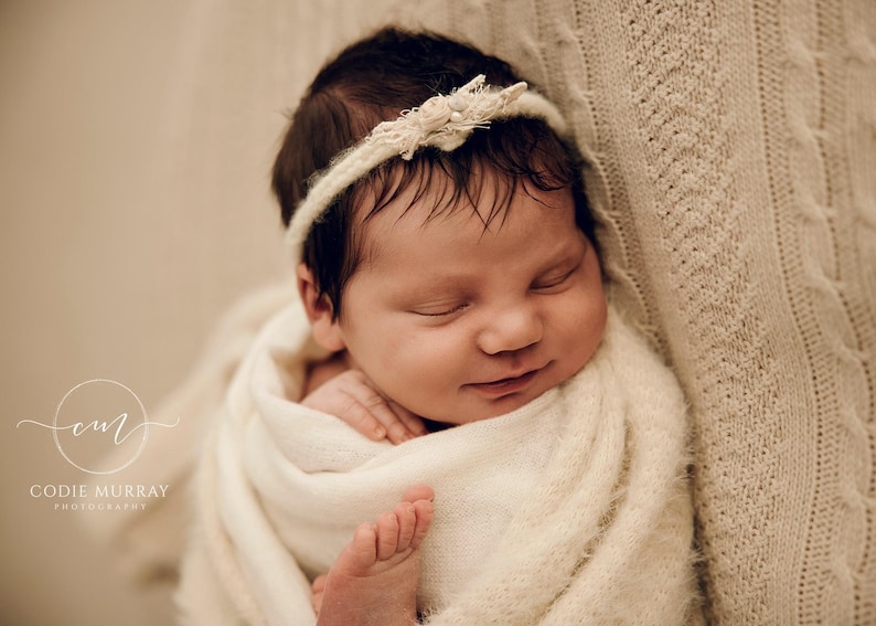 May include: A newborn infant with dark hair is wrapped in a soft, cream-coloured textured blanket. The infant wears a delicate, thin cream-coloured headband adorned with small floral accents. The infant is sleeping peacefully with a slight smile.