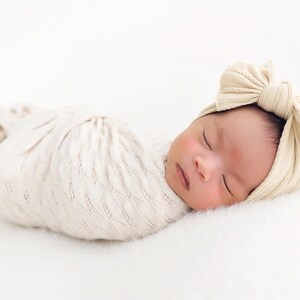 May include: A newborn infant wrapped in a cream-colored swaddle blanket with a textured knit pattern, wearing a matching cream-colored headband featuring a large, prominent bow detail on the side, resting in a peaceful sleeping position.