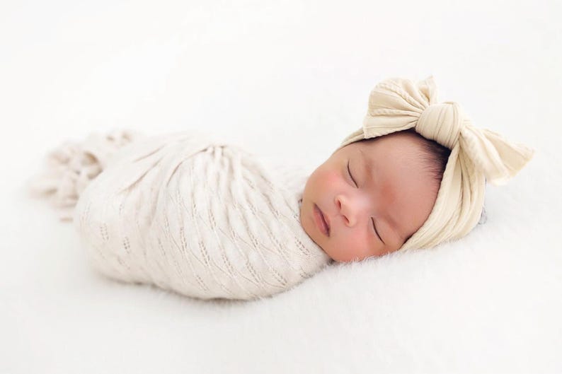 May include: A newborn infant wrapped in a cream-colored swaddle blanket with a textured knit pattern, wearing a matching cream-colored headband featuring a large, prominent bow detail on the side, resting in a peaceful sleeping position.