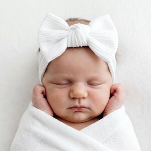 May include: A white ribbed fabric headband featuring a large, centered bow detail. The headband is worn around the head, showcasing a textured, soft material with a knot at the center of the bow, providing a clean and simple aesthetic design.