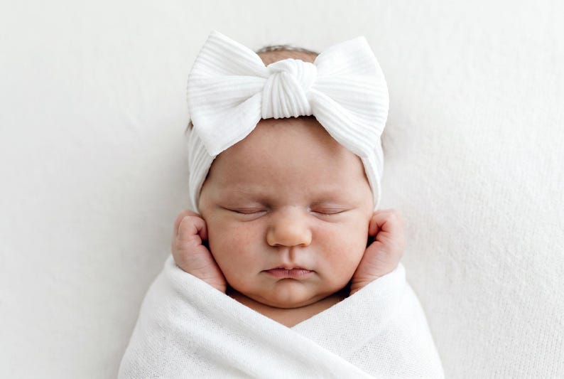 May include: A white ribbed fabric headband featuring a large, centered bow detail. The headband is worn around the head, showcasing a textured, soft material with a knot at the center of the bow, providing a clean and simple aesthetic design.