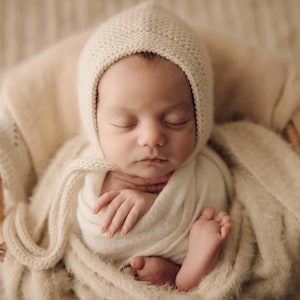 May include: A sleeping newborn infant wrapped in a soft cream-coloured fabric swaddle, wearing a matching cream-coloured knit bonnet with ties, resting inside a woven wicker basket lined with a textured, fuzzy cream-coloured blanket.