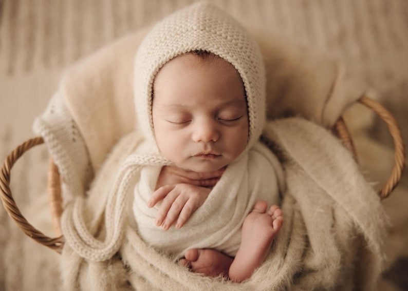 May include: A sleeping newborn infant wrapped in a soft cream-coloured fabric swaddle, wearing a matching cream-coloured knit bonnet with ties, resting inside a woven wicker basket lined with a textured, fuzzy cream-coloured blanket.