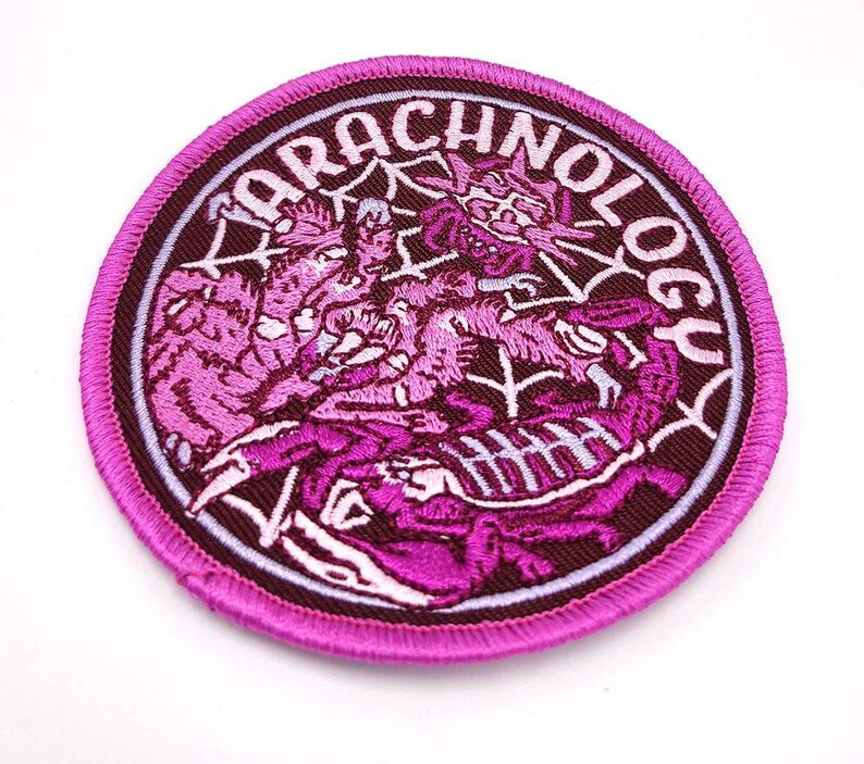 May include: A pink embroidered patch with the word "Arachnology" in a circle around a design of three pink spiders in a web.