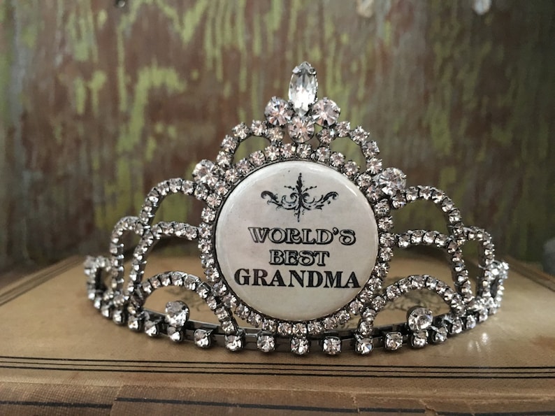 May include: A silver-toned tiara with clear rhinestones and a central circular emblem. The emblem reads "WORLD'S BEST GRANDMA" in black text. The tiara is likely a decorative item, possibly for a special occasion or gift.