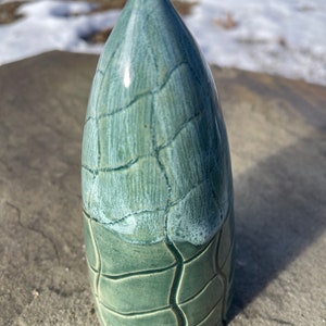 May include: A ceramic sculpture in the shape of a pointed spire with a light green glaze. The glaze has a crackled effect, with a lighter green color at the top and a darker green color at the bottom.