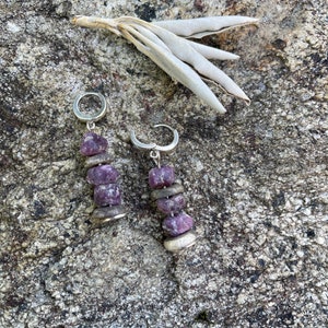 May include: A pair of silver hoop earrings with purple gemstone beads dangling from them. The earrings are on a gray textured surface.