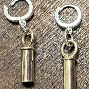 May include: A pair of silver hoop earrings with a gold-colored cylindrical charm hanging from each. The earrings are displayed on a wooden surface. The earrings are a simple, yet elegant design.