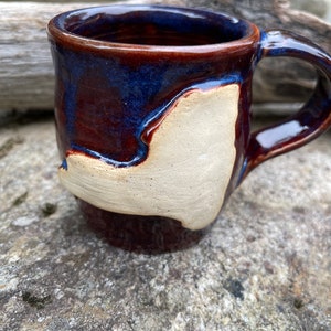 May include: A handmade ceramic mug with a brown, blue, and white glaze. The mug has a unique shape with a white design on the side.