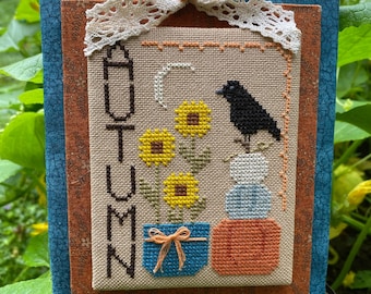 May include: A framed cross-stitch design featuring the word "AUTUMN" in a brown font, with a black crow perched on a stack of white and blue pumpkins. The design also includes sunflowers in a blue pot with a brown ribbon.