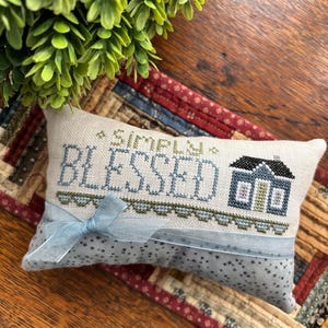 May include: A rectangular, cream-colored decorative pillow with the words "SIMPLY BLESSED" embroidered in blue thread. A small house is stitched next to the word "BLESSED". A light blue ribbon is tied around the pillow. The pillow is on a patterned fabric.