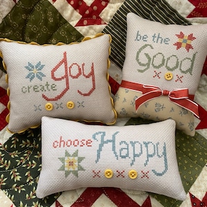 May include: Three white fabric pillow covers with embroidered messages. The first pillow says "create joy" in red and blue lettering. The second pillow says "be the good" in green lettering. The third pillow says "choose happy" in blue lettering. All three pillows have yellow buttons.
