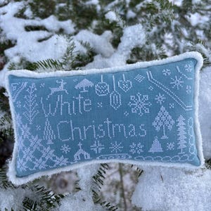 May include: A rectangular, light blue pillow with white embroidered text and winter-themed designs. The text reads "White Christmas" with images of snowflakes, trees, and ornaments. The pillow has a white, fluffy border and is set against a snowy, evergreen backdrop.