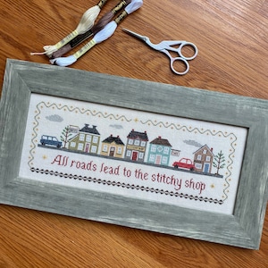May include: A framed cross-stitch design featuring a row of colorful houses with the text "All roads lead to the stitch shop".