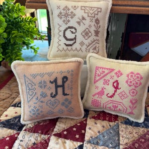 May include: Three square fabric pincushions with white fuzzy trim. The top cushion is cream with brown cross-stitch embroidery featuring the letter G. The bottom left is peach with blue embroidery and the letter H. The bottom right is cream with pink embroidery and the letter J.
