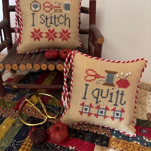 Può includere: Due piccoli cuscini quadrati beige con bordatura in corda rossa e bianca. Uno dice "I stitch" con immagini a punto croce di strumenti di cucito. L'altro dice "I quilt" con immagini simili. Forbici dorate e piccole zucche sono vicine.