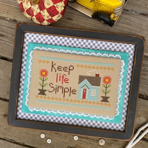 May include: A framed cross-stitch design with the text "Keep life simple" and a small blue house with a red door. The design is surrounded by a white lace trim and a blue and white gingham fabric.