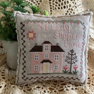 May include: A beige pillow with a brown and pink house design and the text "Till Lady Slipper Lane" embroidered on it.
