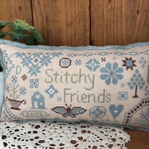 May include: A beige pillow with blue and green embroidery depicting a butterfly, a house, a tree, and flowers. The pillow has a blue fringe and the text "Stitch Friends" is embroidered in the center.