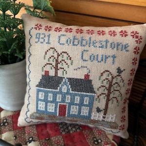 May include: A cross-stitched pillow with a blue house, trees, and the text "931 Cobblestone Court." The pillow has a beige background with a red and blue floral border. The house has a red door and a black roof.