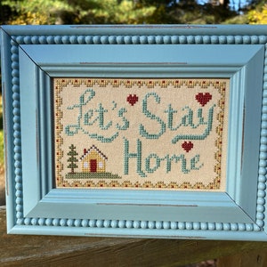Könnte beinhalten: Ein gerahmtes Kreuzstichbild mit einem blauen Rahmen und einem weißen Hintergrund. Der Kreuzstich sagt "Let's Stay Home" mit einem kleinen Haus und Bäumen im Hintergrund.