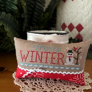 May include: A rectangular fabric pincushion featuring cross-stitch embroidery on tan linen. The text reads "frosty WINTER" in blue and red. A snowman and a red bird on a tree are stitched to the right. The bottom section is red fabric with white patterns.