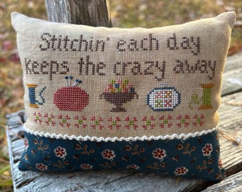 May include: A rectangular, beige pillow with the embroidered text "Stitchin' each day keeps the crazy away." The pillow features cross-stitch designs of sewing tools and a floral border. The bottom is a dark blue fabric with a floral pattern.