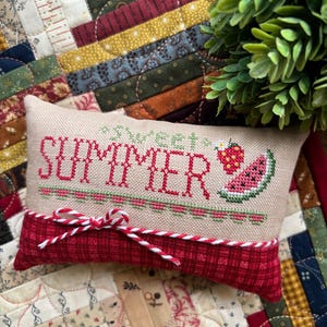 May include: A rectangular, beige fabric pillow with cross-stitched text that reads "A Sweet Summer." The design includes a strawberry and watermelon slice. The bottom of the pillow is red plaid fabric, tied with white and red twine. The pillow is on a patchwork quilt.