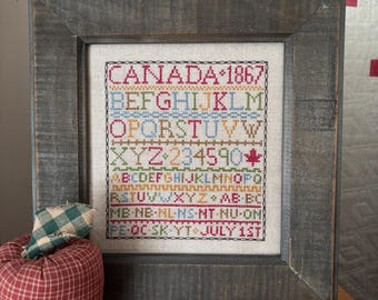 May include: A framed cross-stitch sampler with the text "CANADA 1867" and other letters and numbers in various colors. The frame is a weathered gray wood. An apple-shaped fabric decoration and a small quilted coaster are in the foreground.
