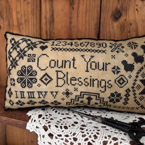 May include: A beige pillow with black embroidery featuring the text "Count Your Blessings" surrounded by various black and white patterns.