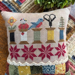 May include: A beige, square decorative cushion with cross-stitch embroidery. The design features spools of thread, a bird, scissors, and floral elements. The bottom edge has a patchwork design with a scalloped trim.