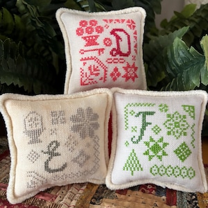 May include: Three small, square, off-white decorative pillows with embroidered designs. One pillow features a red "D", another has a gray "E", and the third displays a green "F". Each pillow has a fluffy, cream-colored border.