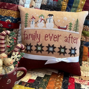 May include: A decorative pillow with a cross-stitched design featuring snowmen, trees, and the phrase "family ever after." The pillow has a tan background with a dark red bottom and a white ribbon. The pillow is approximately 8 inches wide and 6 inches tall.
