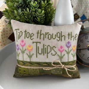 May include: A beige cushion with a green embroidered design of tulips and the text "Tiptoe through the Tulips". The cushion is decorated with a brown twine bow and a green fabric backing.