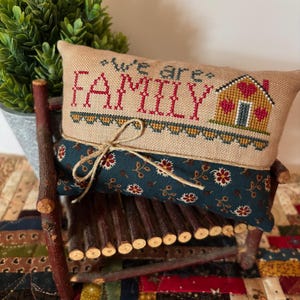 Peut inclure: Un coussin décoratif brodé avec les mots "we are FAMILY" en fil rouge. Il présente un motif de maison et repose sur une petite chaise en bois. Le coussin est orné d'un tissu à motifs floraux sur le bas.