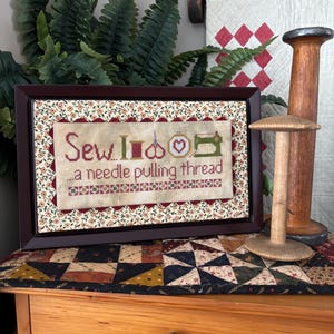 Puede incluir: Arte de punto de cruz enmarcado con las palabras "Sew I do... a needle pulling thread" e iconos de costura. El marco es marrón oscuro y la obra presenta colores rojo, verde y crema. Un carrete de madera y un portahilos están a la derecha.
