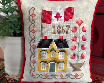 May include: A white pillow with a red border and a cross-stitch design featuring a yellow house, a red poppy flower, and a Canadian flag with the year 1867.