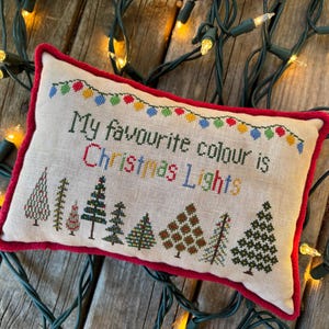 May include: A rectangular, off-white pillow with red trim features cross-stitched text that reads "My favourite colour is Christmas Lights." A string of colorful lights is stitched above the text. Below the text are cross-stitched Christmas trees. The pillow is surrounded by a string of Christmas lights.
