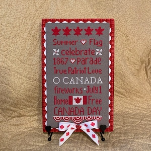 May include: A gray and red cross-stitch sampler with a Canadian theme. The sampler features the text "Summer Flag * Celebrate * 1867 Parade True Patriot Love O CANADA fireworks July 1 Home Free CANADA DAY".