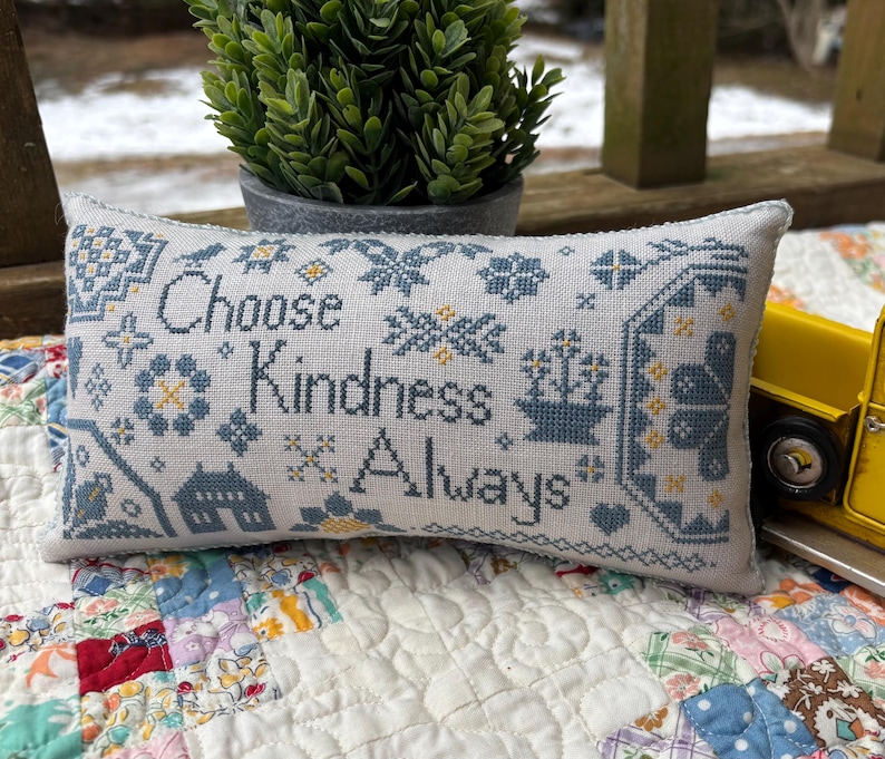 May include: A white pillow with blue and yellow embroidery. The pillow features the text "Choose Kindness Always".