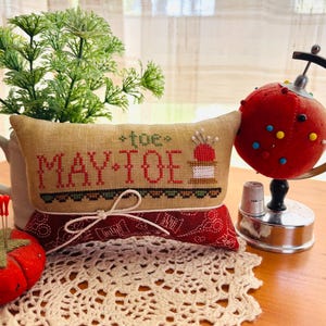 Può includere: Un cuscino per spilli fatto a mano con la scritta "MAY TOE" ricamata su tessuto beige. Il cuscino poggia su un tessuto rosso fantasia e un centrino color crema. Sono visibili anche un portaspilli a forma di pomodoro e un portaspilli a forma di globo rosso.