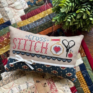 May include: A rectangular cross-stitch pillow with the words "Cross Stitch" embroidered in red. The pillow features a heart, scissors, and a needle and thread design. A white ribbon is tied on the bottom. The pillow is on a patchwork quilt.