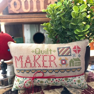 May include: A rectangular beige pincushion with the words "Quilt Maker" embroidered in red and green thread. The bottom half features a floral pattern, and a red string is tied around the middle. The pincushion rests on a patchwork quilt.