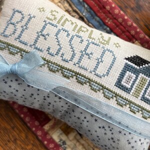 K&ouml;nnte beinhalten: Ein rechteckiger, cremefarbener Stoffartikel mit den Worten "SIMPLY BLESSED" in Blau und Gr&uuml;n gekreuzt. Auf der rechten Seite ist ein kleines Haus gestickt. Ein hellblaues Band ist um den Artikel gebunden. Der Stoff hat ein Muster aus kleinen blauen Sternen.