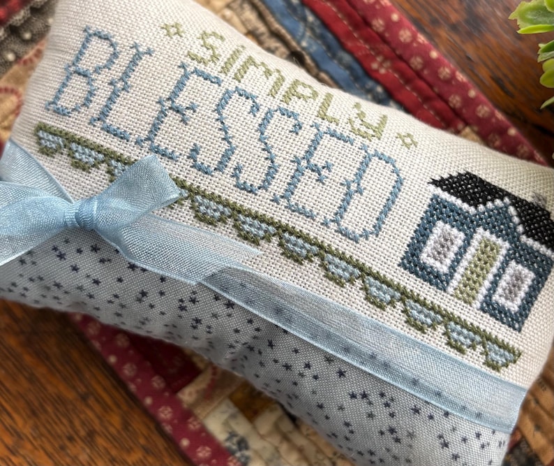 K&ouml;nnte beinhalten: Ein rechteckiger, cremefarbener Stoffartikel mit den Worten "SIMPLY BLESSED" in Blau und Gr&uuml;n gekreuzt. Auf der rechten Seite ist ein kleines Haus gestickt. Ein hellblaues Band ist um den Artikel gebunden. Der Stoff hat ein Muster aus kleinen blauen Sternen.