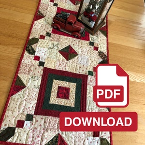 May include: A red, green and white quilted table runner with a Christmas theme. The runner features a central square with a smaller square inside, surrounded by a border of green and red squares. The runner is decorated with a red truck carrying a small Christmas tree, and a glass lantern with a snowy scene inside. The text "PDF DOWNLOAD" is visible in the lower right corner of the image.