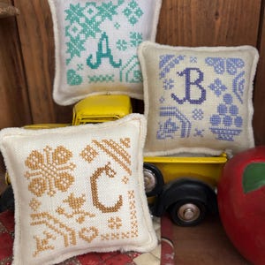 May include: Three small, square, embroidered pillows with the letters A, B, and C. The pillows feature cross-stitch designs in teal, purple, and gold, with decorative patterns. The pillows are on a wooden surface with a yellow toy truck.