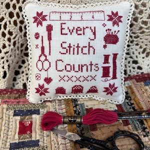May include: A white pillow with red embroidery that reads "Every Stitch Counts". The pillow is surrounded by a white crocheted border. The pillow is sitting on a patchwork quilt with a red and white embroidery floss and a pair of black scissors.