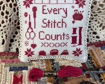 May include: A white pillow with red embroidery that reads "Every Stitch Counts". The pillow is surrounded by a white crocheted border. The pillow is sitting on a patchwork quilt with a red and white embroidery floss and a pair of black scissors.
