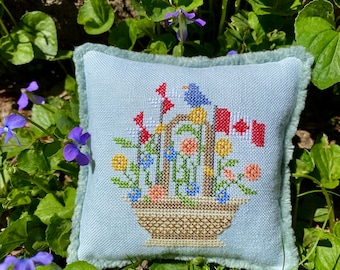 May include: A blue and white embroidered pillow with a floral design and a bird in a basket. The pillow has a light blue background and a white border.